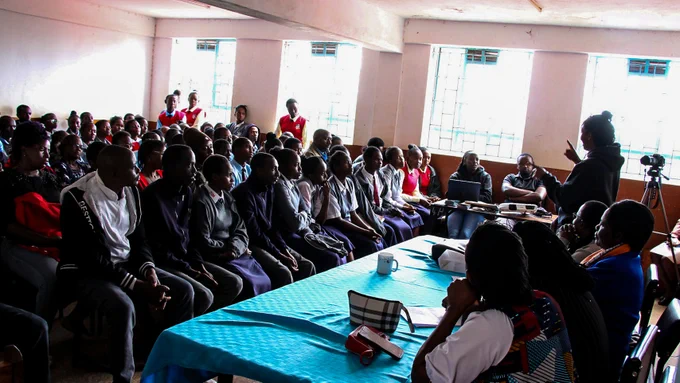A facilitator stands at the front of the room addressing the audience, using hand gestures to emphasize key points. The audience consists mainly of secondary school students, many wearing school uniforms, seated closely together and attentively listening. Several adults, teachers, and parents are also present among the participants.