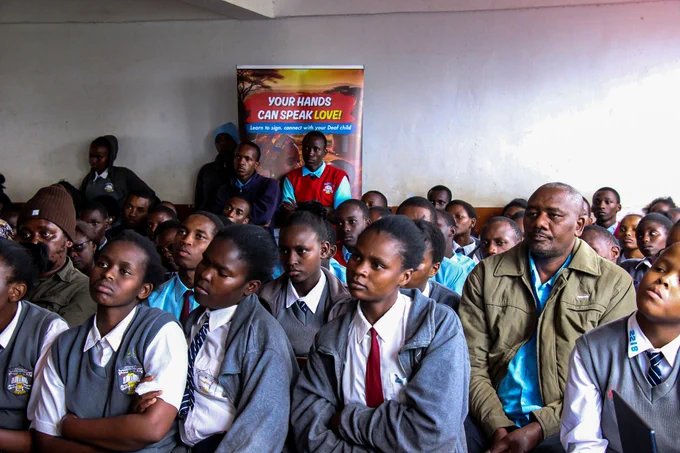 The image is a group of people, predominantly young students, seated indoors. The main focus is on the students in the foreground, most of whom are wearing school uniforms consisting of grey vests over white shirts, some with ties. The overall atmosphere appears to be one of attention and engagement, as many individuals are looking forward at a speaker.In the center of the background, a colorful banner is visible with the text "YOUR HANDS CAN SPEAK LOVE!" and "Learn to sign, connect with your Deaf child".