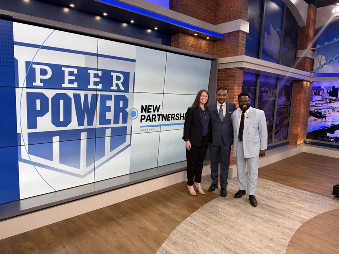 Kate, Cochran, Alex Coleman, and Cortney Richardson stand side by side in the news room, smiling at the camera. A screen next to them reads "Peer Power - New Partnership"