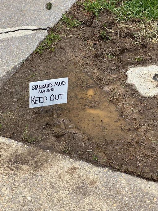 A patch of mud with a small sign reading "Standard Mud, SRM 7581, Keep Out" is situated next to a cracked sidewalk and grass.