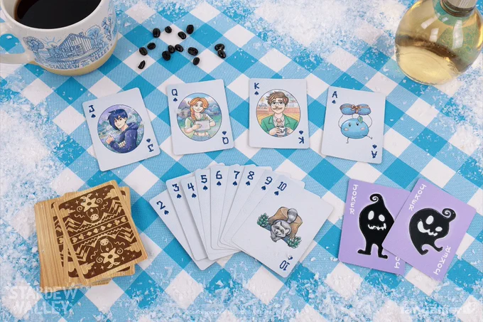 A photo of the Spades from the Stardew Valley playing card deck against a gingham picnic tablecloth, showing a wintery theme and two Jokers with Krobus on the face. 