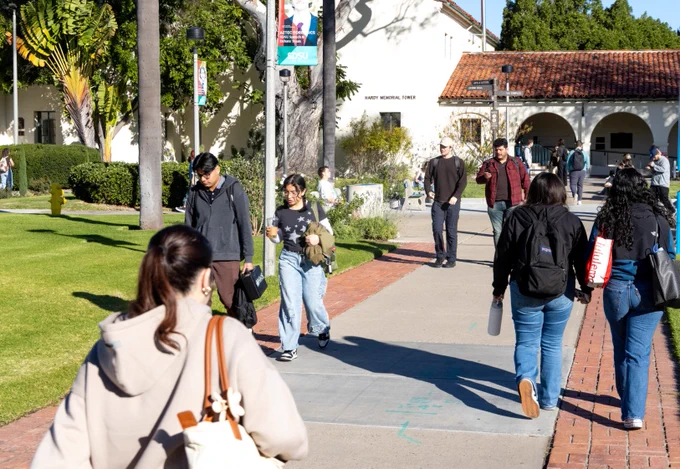 People walking on a sunny pathway at a college campus, with a building labeled "Hardy Memorial Tower" in the background. Some individuals are carrying backpacks and passing by green lawns and palm trees.