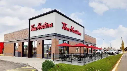 Exterior of a Tim Hortons cafe with red signage, brick facade, outdoor seating under red umbrellas, clear sky, and a grassy pathway.