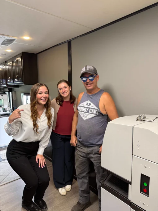 Three people posing inside a modern RV kitchen area with dark cabinets and a large white appliance nearby.