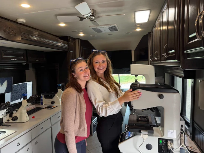 Two women standing inside a mobile lab, one operating a large microscope while the other observes nearby.