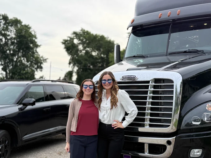 Two women standing side by side in front of a large black semi-truck and a black SUV parked outdoors with trees in the background.