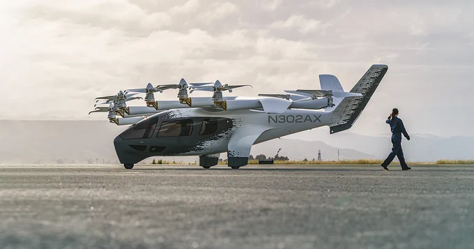 A futuristic aircraft with multiple rotors is parked on a runway. A person is walking nearby, holding something to their ear, possibly a phone. The background features a hazy sky and distant hills.