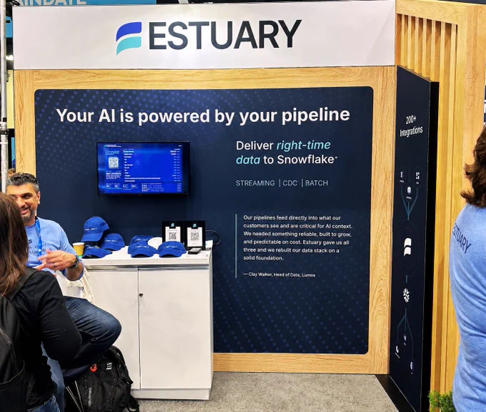 Estuary booth at a trade show with a display featuring a message about delivering real-time data to Snowflake. A monitor, promotional materials, and branded items like hats are on a table. A person stands nearby, partially visible.