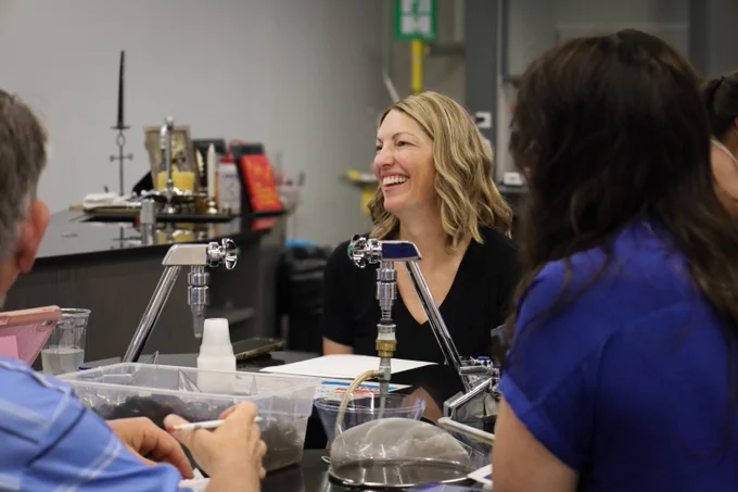 Three people engaged in a discussion around a lab table with scientific equipment and supplies in a classroom setting.
