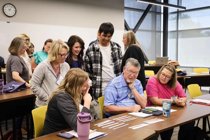 Group of adults engaged in a collaborative activity around a table in a bright classroom with large windows and yellow chairs.
