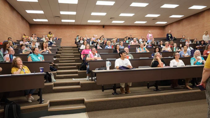 A diverse group of adults attentively listening in a large university lecture hall with tiered seating and bright overhead lights.