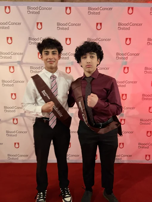 Two young men stand on a red carpet at a Blood Cancer United event, wearing candidate sashes and formal attire. The mood is formal and supportive.