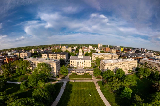 Aerial view of a university campus with a central historic building featuring a clock tower surrounded by green lawns and trees under a partly cloudy sky.