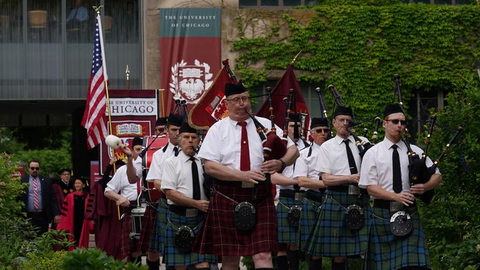 A marching band parades, led by bagpipers in kilts, followed by drummers, in front of \"THE UNIVERSITY OF CHICAGO\" banners. An American flag is seen alongside people in graduation gowns.