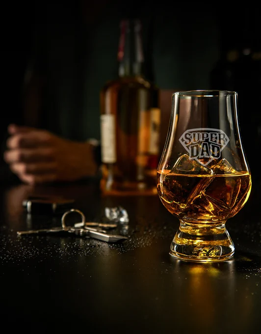 Engraved "Super Dad" whiskey glass with amber-colored liquid and ice on a dark tabletop. A bottle, keys, and a person are blurred in the background.