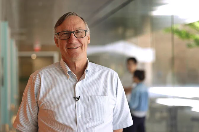 Physicist Stephen Forrest smiling and wearing glasses and a striped short-sleeved shirt standing in a modern hallway.