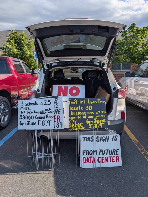 Signs in the trunk of a car "3 schools in 25 miles. Tell Lyon Twp no data center. 58000 Grand River 6pm June 1, 8, 9" 