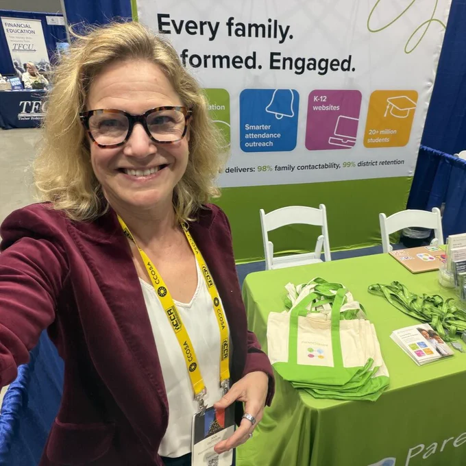 Person taking a selfie at an educational conference booth. The booth displays a banner with text promoting family engagement, smarter attendance outreach, and K-12 websites. Some educational materials and tote bags are arranged on a table.
