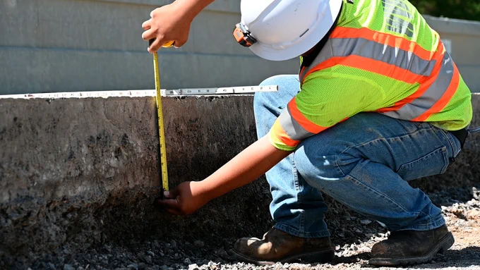 A person wearing a white hard hat and a high-visibility safety vest is crouching on a construction site. They are using a yellow measuring tape to check the height of a concrete barrier. The person is on a gravel surface, and part of the structure is visible.