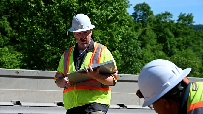 Two construction workers wearing high-visibility vests and hard hats are on a site. One is holding a clipboard, and the scene is set against a backdrop of greenery.