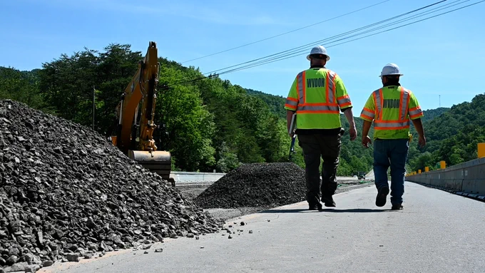 Two individuals wearing high-visibility vests labeled "WVDOH" walk on a road under construction. A large pile of stone is on one side, and an excavator is in the background. Trees and a clear blue sky are visible.