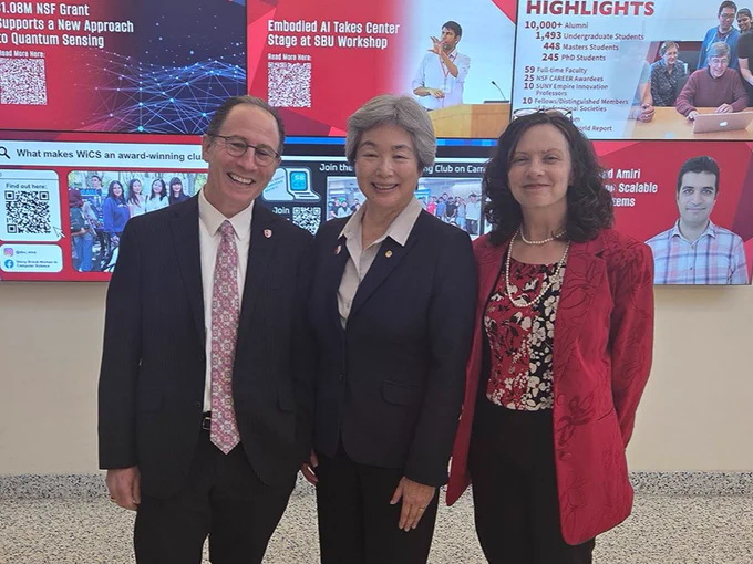 A group of three people posing in front of a large digital display featuring various Stony Brook University highlights and news. The content includes information on quantum sensing, AI workshops, and student achievements. They are standing in a spacious lobby area.