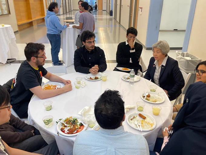 A group of people sits around a circular table with plates of food and drinks. They appear to be engaged in conversation. In the background, a few more individuals stand near another table in a hallway. The setting suggests an informal gathering or event.