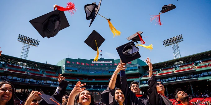Graduates celebrate at Fenway Park, tossing their caps into the air under a clear blue sky. The iconic park's seating and lights are visible in the background.
