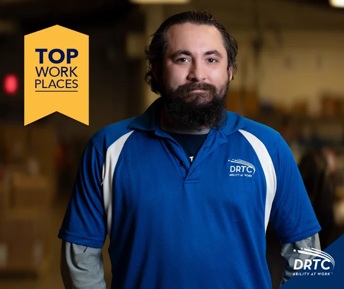 Employee wearing a blue DRTC polo shirt standing in a warehouse environment with a Top Work Places banner.