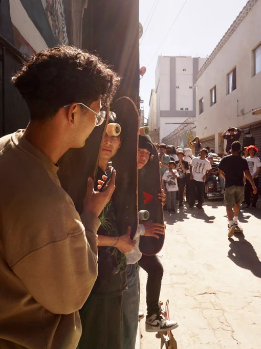 A group of people stand in an alley holding skateboards, with a crowd of skateboarders in the background. Sunlight streams between the buildings.