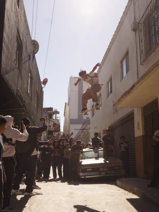 A person is skateboarding in mid-air above an alley, performing a trick. A group of onlookers gathers below, some holding cameras. The alley is flanked by tall buildings, and a parked car is visible. The scene is lively and captures a sense of action.