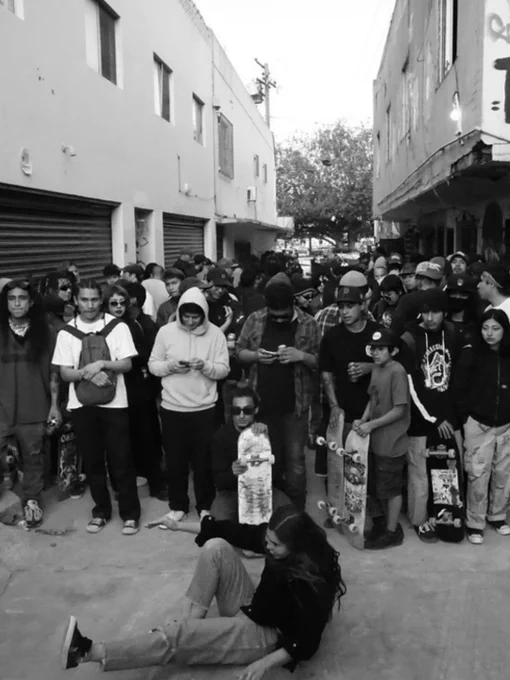 A large group of people, many holding skateboards, are gathered in an alleyway between two buildings. The crowd includes individuals standing and some sitting on the ground. The setting appears casual and urban, with closed storefronts visible in the background.