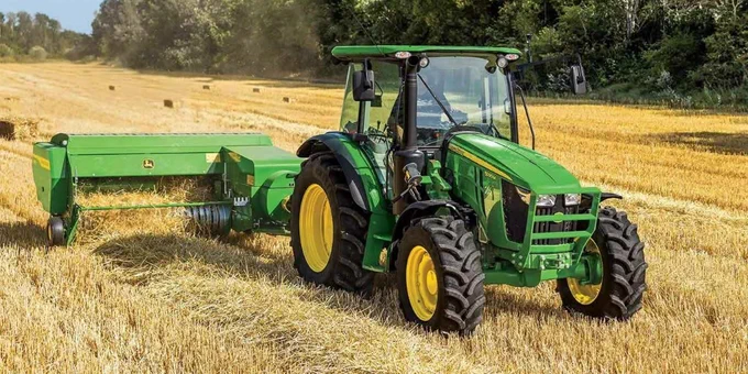 A green John Deere tractor is working in a golden field, pulling a piece of agricultural equipment that appears to be for harvesting. The background features trees and a clear sky.