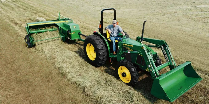 A person operates a green John Deere tractor with a hay rake attachment, working in a field of cut hay.
