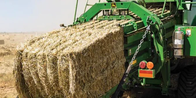 A large green John Deere baler is ejecting a rectangular bale of hay onto a dry, grassy field. The machinery is detailed with several metal components and safety markings.