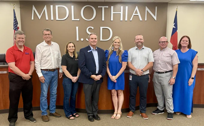 A group of eight people standing in front of a sign that reads "Midlothian I.S.D." with two flags visible on either side. They are all smiling and dressed in business or casual attire.