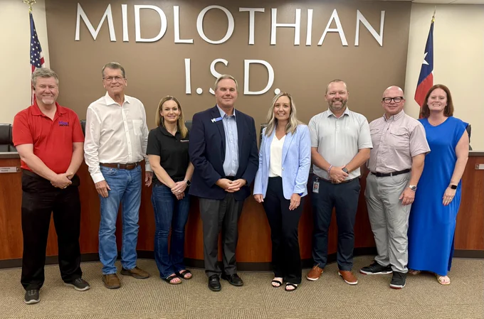 Eight individuals are standing in front of a wall with the text "MIDLOTHIAN I.S.D." There are two flags on either side of the group. They are inside a room with a wood-paneled lectern.