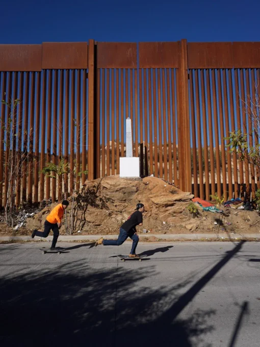 Two people skateboard on a sunny day in front of the US-Mexico border wall. The rust-colored metal bars of the wall rise tall in the background, and there's a small obelisk monument on a rocky mound between them. Sparse vegetation is visible around the rocky area.
