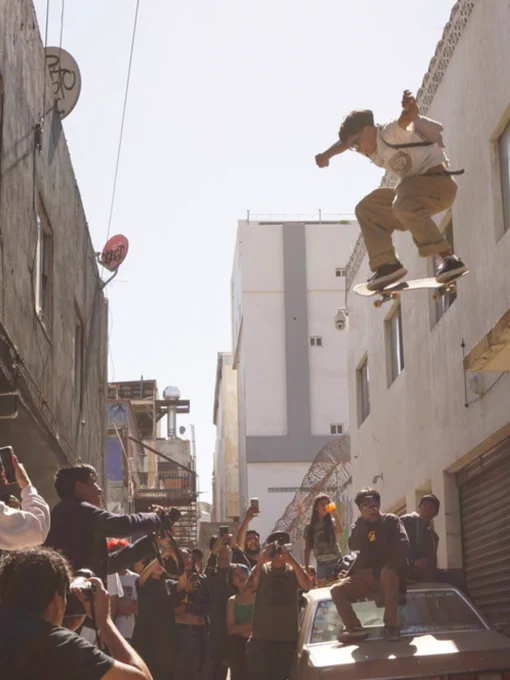 A person skateboards off a ledge in a narrow alley, surrounded by spectators taking photos and videos. The scene captures an urban environment with tall buildings and utility wires. The onlookers stand on the ground and atop a vehicle, watching the skateboarder mid-air.