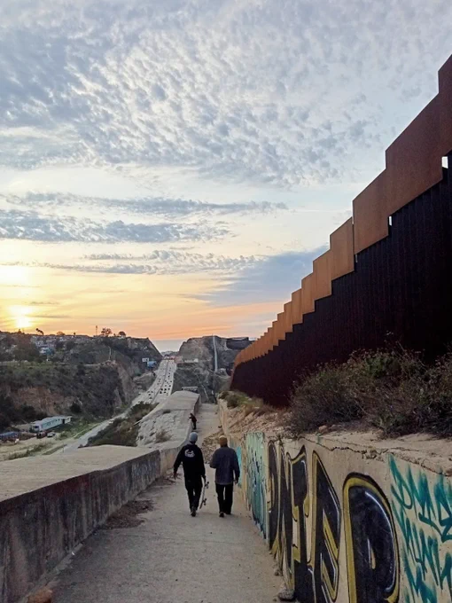 A walkway beside a tall border fence stretches into the distance. Two individuals walk along the path, which is lined with graffiti-covered walls. The sky is filled with scattered clouds, illuminated by the setting sun.