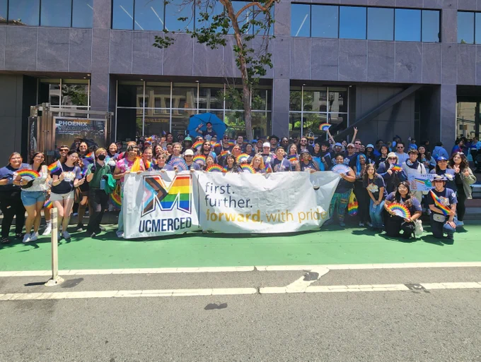 A group of UC Merced students, staff, faculty, and alumni holding a large "UC Merced" banner with a multi-colored "M" and the slogan "first. further. forward. with pride" taken in 2024.