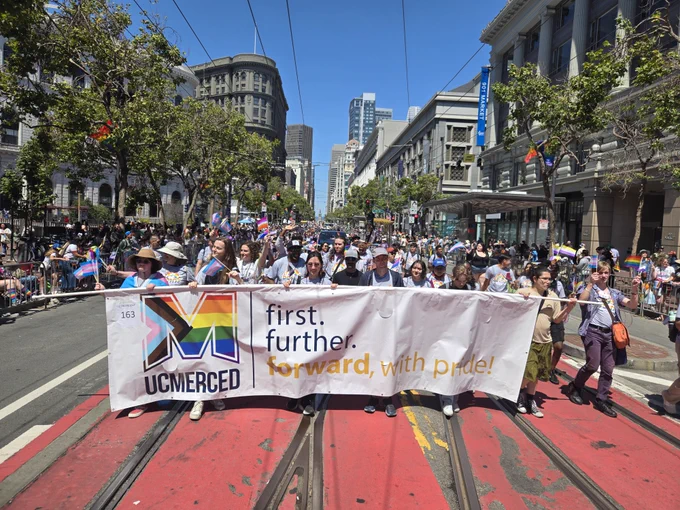 A group of UC Merced students, staff, faculty, and alumni holding a large "UC Merced" banner with a multi-colored "M" and the slogan "first. further. forward. with pride" taken in 2025 as they walk the San Francisco Pride Parade.