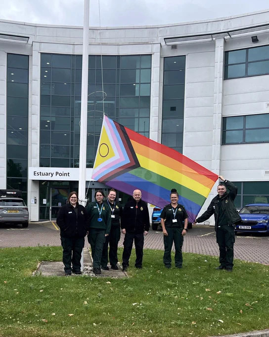 A group of NWAS colleagues stand outside Estuary Point beside a large Progress Pride flag during a flag-raising event.