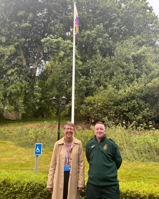 Two colleagues stand in front of a flagpole with the Progress Pride flag flying at an NWAS site.