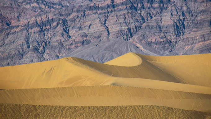 Dunes at Mesquite Flat in Death Valley National Park, with smooth golden curves in the foreground and rugged, multicolored mountains in the distance under clear desert light.