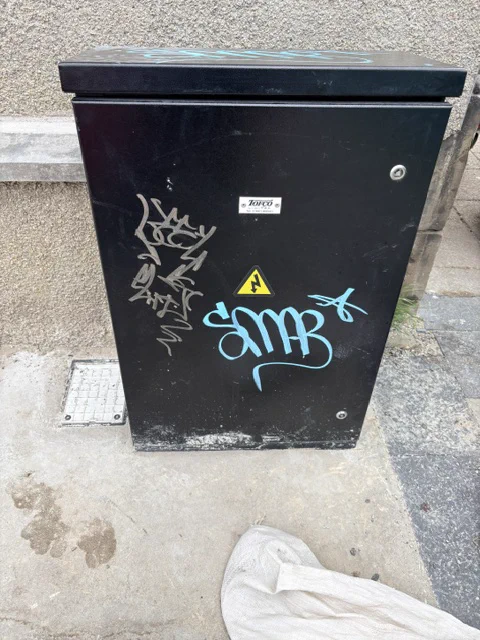 A black street cabinet covered in graffiti, featuring a yellow lightning bolt warning sign.