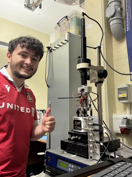 Person in a red United shirt giving thumbs up next to a complex laboratory instrument with wires and control panels in a research setting.