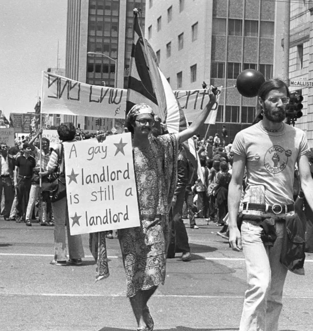Man in a parade holding a sign that says "A gay landlord is still a landlord." Another man in front of him is wearing a "faggot revolution" t-shirt.
