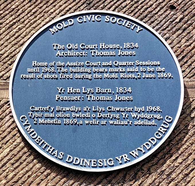 A blue plaque by Mold Civic Society on the wall of the Old Court House, 1834, with the following words: Home of the Assize Court and Quarter Sessions until 1968. The building bears marks said to be the result of shots fired during the Mold Riots, 2 June 1869.