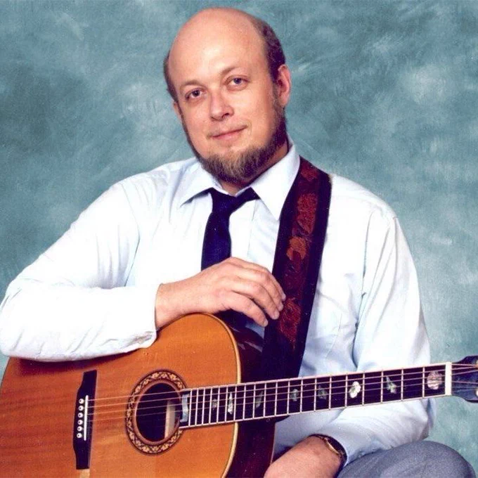 Stan Rogers sits for a studio portrait holding an acoustic guitar across his lap. He wears a light blue dress shirt with a dark tie and patterned guitar strap. He has a bald head, short beard, and slight smile, with a mottled blue-gray backdrop behind him.
